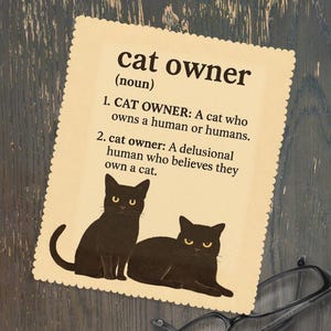 Puede incluir: Una tarjeta beige con la definición de "cat owner" y dos ilustraciones de gatos negros. El texto dice: "CAT OWNER: Un gato que posee a un humano o humanos. cat owner: Un humano delirante que cree que posee un gato."