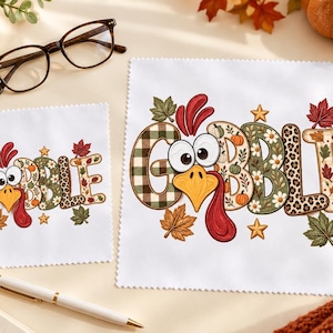 May include: Two white square microfiber cloths featuring a colorful graphic of a turkey face integrated into the word GOBBLE. The letters are decorated with patterns including green plaid, pumpkins, flowers, and leopard print, surrounded by autumn leaves.