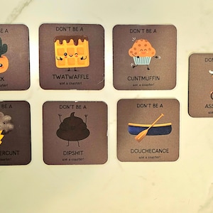 May include: Eight square coasters with humorous illustrations and text. Each coaster features a different cartoon character and a rude or offensive word or phrase. The coasters are brown with white text and illustrations.