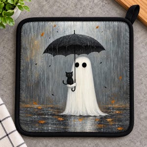 May include: A square pot holder featuring a white ghost holding a black umbrella over a small black cat. The scene depicts a rainy day with falling autumn leaves against a grey background, finished with a black fabric border and a hanging loop.