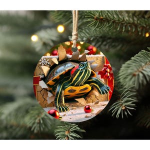 May include: A round Christmas ornament featuring a green, black, and yellow turtle peeking out of a ripped, brown gift box with red and white accents. The turtle has a red stripe on its shell.