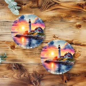 May include: Two round coasters with a watercolor painting of a lighthouse at sunset. The lighthouse is white with a red roof and is surrounded by water and rocks. The sky is a mix of pink, orange, and purple.