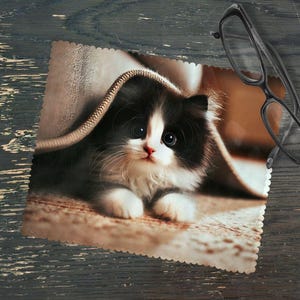 Pode incluir: Um gatinho preto e branco com olhos verdes espreita por baixo de um cobertor castanho. O gatinho está deitado numa superfície castanho claro.