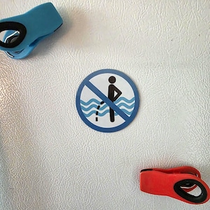 May include: A white refrigerator with a blue and a red clip, and a round sign with a blue border. The sign has a black figure urinating in water with a diagonal red line through it, indicating a prohibition.