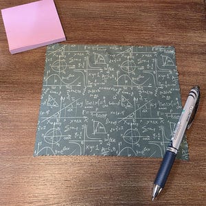 May include: A green cloth with white mathematical equations and diagrams, a pink sticky note pad, and a blue and silver pen with the text "Metal Tip 07" on a wooden surface. The cloth has a frayed edge.
