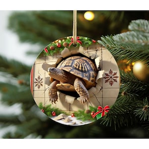 May include: A round ceramic Christmas ornament featuring a brown and tan tortoise emerging from a hole in a tiled wall. The design includes green pine branches, red bows, and brown snowflake patterns on a light-colored background with a gold string.