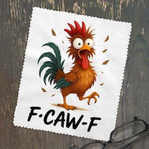 May include: White cleaning cloth with a cartoon rooster illustration. The rooster is brown, red, and green with a surprised expression and its tongue sticking out. The text "F.CAW-F" is printed in black. A pair of glasses rests on the wooden surface.