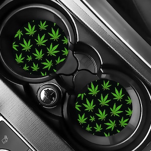 May include: Black car coasters featuring a repeating pattern of green cannabis leaves. The coasters are circular and designed to fit into car cup holders. The car's interior is black and silver.