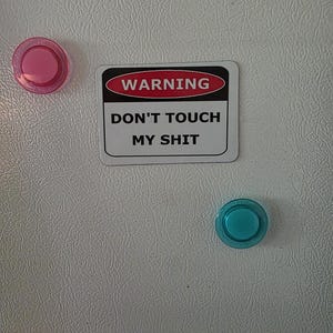 May include: A rectangular magnet with the text "WARNING DON'T TOUCH MY SHIT" in black and white, with a red and black warning symbol. Two round magnets, one pink and one teal, are also visible.