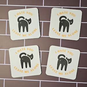 May include: Four square coasters with a black cat illustration and the text "I hate you leave me alone".
