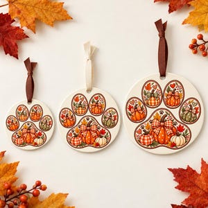 May include: Three round white ceramic ornaments of varying sizes, each featuring a paw print design filled with illustrations of orange, white, and green pumpkins, autumn leaves, and small hearts, suspended by brown or cream-colored ribbons.