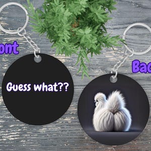 May include: A black round keychain with the text "Guess what?" on the front and a white fluffy chicken on the back.