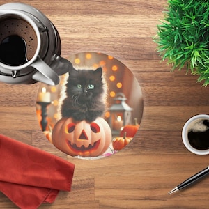 Può includere: Un gattino nero con gli occhi verdi è seduto su una zucca intagliata, parte di un sottobicchiere a tema Halloween. Una caffettiera bianca e una tazza di caffè sono su un tavolo di legno, insieme a un tovagliolo rosso, una penna e una piccola pianta verde.