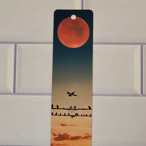 Op de afbeelding: Een bladwijzer met een rode maan in een zonsondergangshemel met vogels die op een draad zitten. De maan is fel oranje en de hemel is een gradiënt van oranje, geel en blauw.