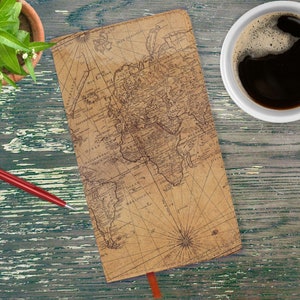 May include: A vintage-style world map notebook with a brown leather cover and a red ribbon bookmark. The notebook is on a wooden table with a cup of coffee and a potted plant.