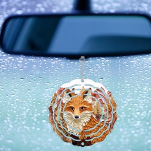 May include: A car air freshener featuring a detailed fox design. The fox has orange and white fur, surrounded by a circular pattern with a light blue outline. The air freshener hangs from a string, with a car's rearview mirror in the background.