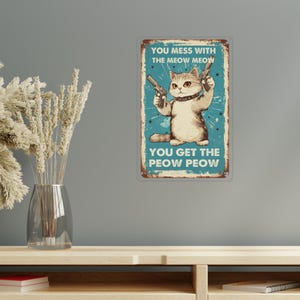 Può includere: Un cartello metallico rettangolare dall'aspetto vintage e usurato. Il cartello presenta un gatto dei cartoni animati che tiene due pistole, con il testo "YOU MESS WITH THE MEOW MEOW, YOU GET THE PEOW PEOW" in un carattere giocoso. Lo sfondo è verde acqua.