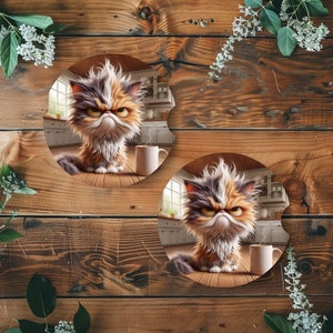 May include: Two round coasters featuring a cartoon image of a grumpy cat with orange, white, and gray fur. The cat is sitting in a kitchen setting with a steaming mug of coffee in front of it. The coasters are made of a cork-like material.
