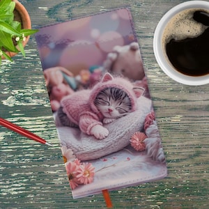Puede incluir: Un diario rosa y blanco con un lindo gatito durmiendo que lleva un gorro rosa en la portada. El gatito está acostado en una manta suave y peluda con flores rosas.