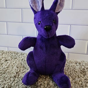 Puede incluir: Un juguete de peluche de canguro morado con una oreja interior de color morado claro. El canguro está sentado en una alfombra de pelo marrón.