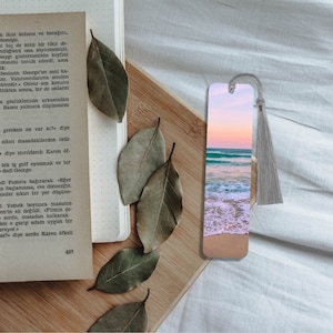 May include: A colorful bookmark with a beach scene featuring a sandy beach, blue ocean waves, and a pink and orange sunset sky. The bookmark has a gray tassel.