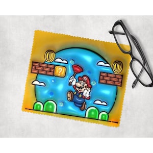 Puede incluir: Un paño de microfibra amarillo con una imagen de dibujos animados de Mario del videojuego Super Mario Bros. Mario está saltando sobre un bloque de ladrillo con un signo de interrogación y una moneda. El fondo es un cielo azul con nubes.