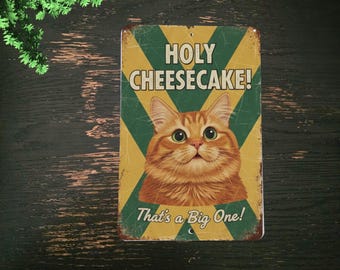 May include: A rectangular metal sign with a distressed finish. The sign features a cartoon image of an orange tabby cat with green eyes. Text on the sign reads "HOLY CHEESECAKE!" and "That's a Big One!" in a playful font, set against a yellow and green background.