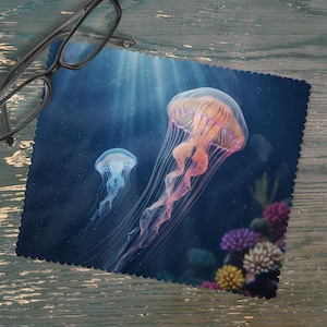 May include: A microfiber cleaning cloth featuring a vibrant underwater scene with two jellyfish. The larger jellyfish is orange and pink, while the smaller one is blue. The cloth has a scalloped edge and is designed for cleaning eyeglasses.
