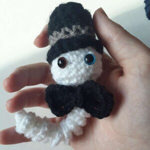 May include: White crocheted amigurumi figure wearing a black top hat with a brown band, a black bow tie, and blue and brown eyes.