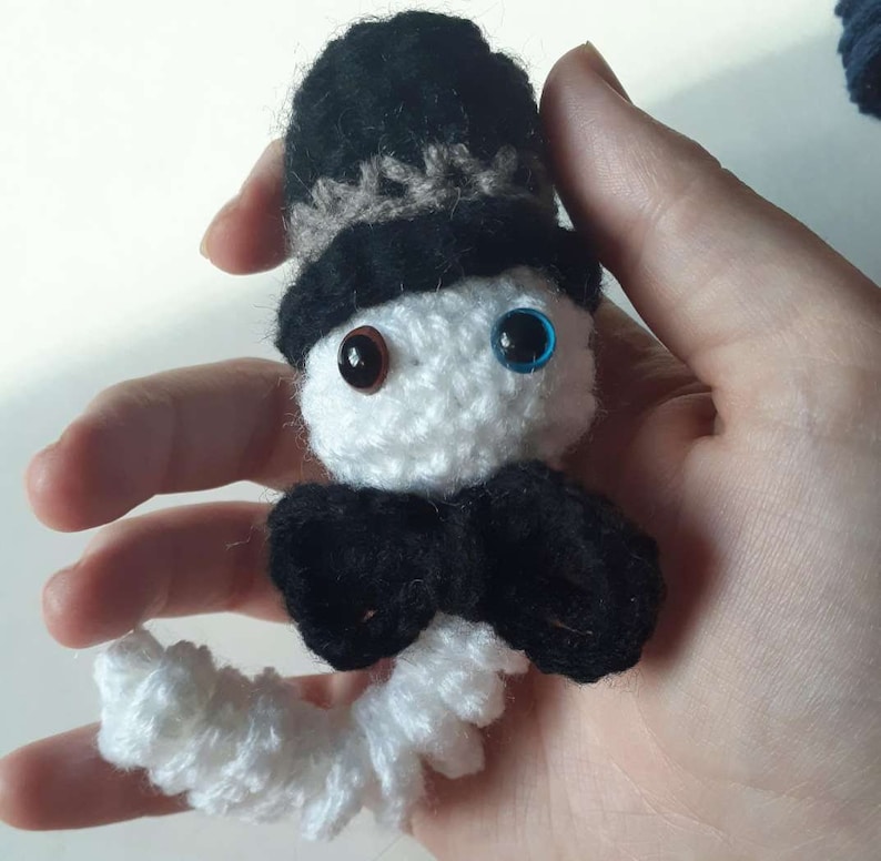May include: White crocheted amigurumi figure wearing a black top hat with a brown band, a black bow tie, and blue and brown eyes.