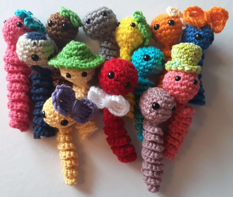 May include: A collection of colourful crocheted worm toys. The worms are made in a variety of colours, including pink, red, yellow, blue, green, brown, and grey. Some of the worms have hats or bows.
