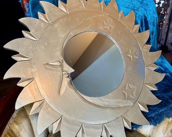 May include: A decorative wall mirror in the shape of a sun and moon. The sun has pointed rays and a face, with a crescent moon and stars. The mirror has a gold finish and is likely made of metal.