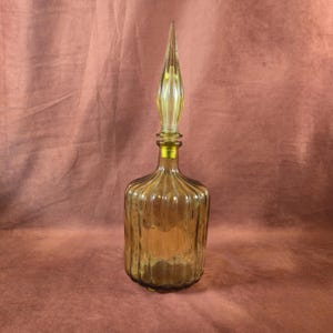 May include: Amber-colored glass bottle with a tall, pointed stopper. The bottle has vertical ribbed detailing and a rounded base. The stopper is a lighter shade of yellow and tapers to a sharp point. A decorative piece.