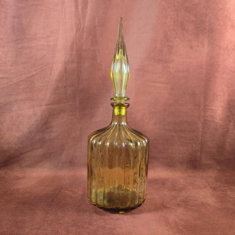 May include: A vintage amber glass bottle with a ribbed design and a tall, pointed stopper. The bottle has a rounded base and a slender neck, with a yellow-tinted stopper. The overall design is elegant and decorative.