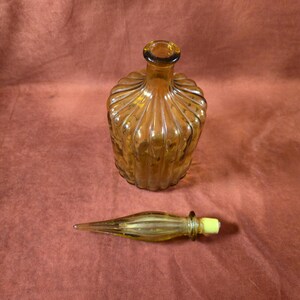 May include: Amber-colored glass bottle with a ribbed design and a separate stopper. The bottle has a rounded shape with a narrow neck and a dark rim. The stopper is elongated with a yellow tip. The bottle and stopper are on a reddish-brown background.