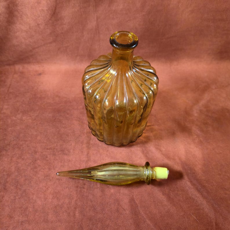 May include: Amber-colored glass bottle with a ribbed design and a separate stopper. The bottle has a rounded shape with a narrow neck and a dark rim. The stopper is elongated with a yellow tip. The bottle and stopper are on a reddish-brown background.