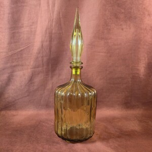 May include: Amber-colored glass decanter with a tall, pointed stopper. The bottle has vertical ribbed detailing and a rounded base. The stopper is a lighter shade of amber and has a long, slender shape. The decanter is likely used for storing and serving beverages.