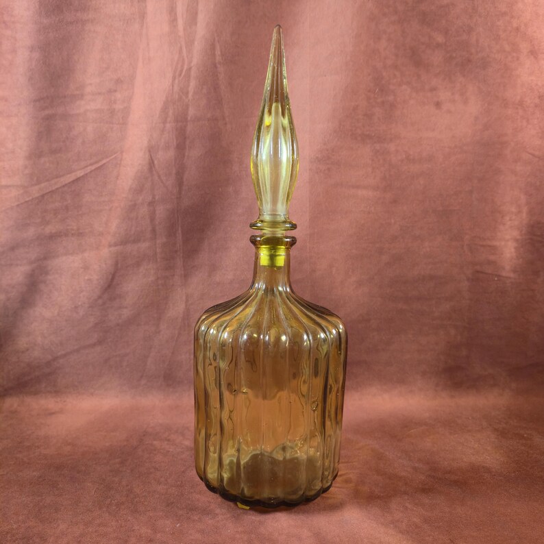 May include: Amber-colored glass decanter with a tall, pointed stopper. The bottle has vertical ribbed detailing and a rounded base. The stopper is a lighter shade of amber and has a long, slender shape. The decanter is likely used for storing and serving beverages.