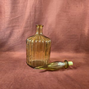 May include: Amber glass bottle with a ribbed design and a matching stopper. The bottle has a rounded shape and a long neck. The stopper is lying next to the bottle. The items are on a reddish-brown surface.
