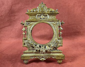 May include: Ornate, antique-style gold-colored picture frame with a circular opening. The frame features detailed floral and scrollwork designs, with decorative elements on the sides and top. The frame is freestanding.