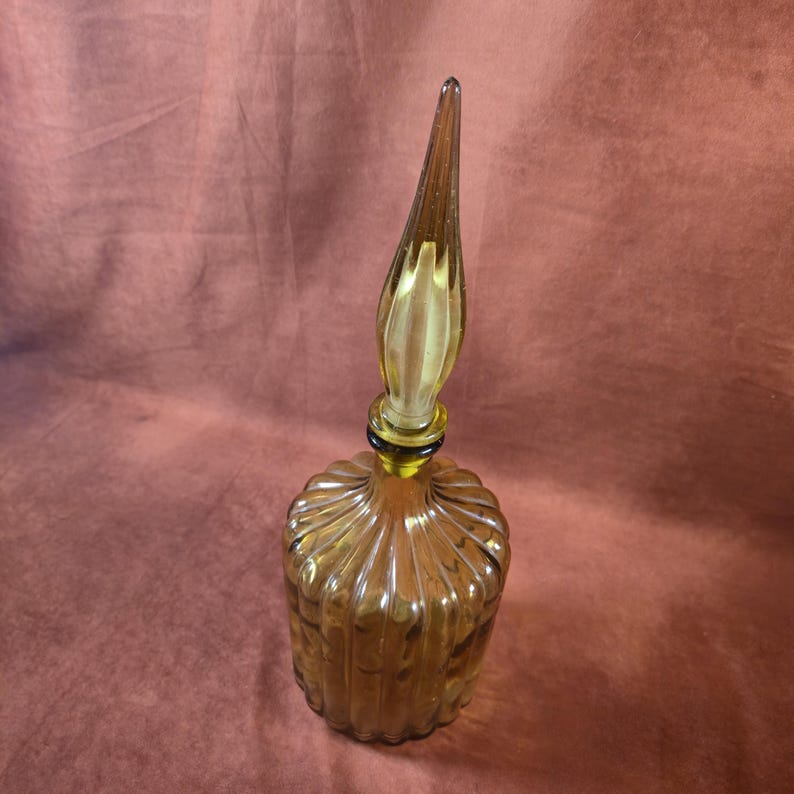 May include: Amber-colored glass bottle with a ribbed, rounded body and a tall, pointed stopper. The bottle has a vintage aesthetic, with a classic design. The stopper is a lighter shade of amber, adding contrast to the overall look. The bottle is likely used for perfume or decorative purposes.