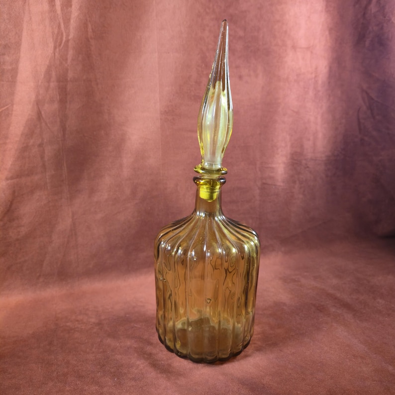 May include: Amber-colored glass bottle with a tall, pointed stopper. The bottle has vertical ribbed detailing and a rounded base. The stopper is a lighter shade of amber and tapers to a sharp point. The bottle is likely for decorative use.