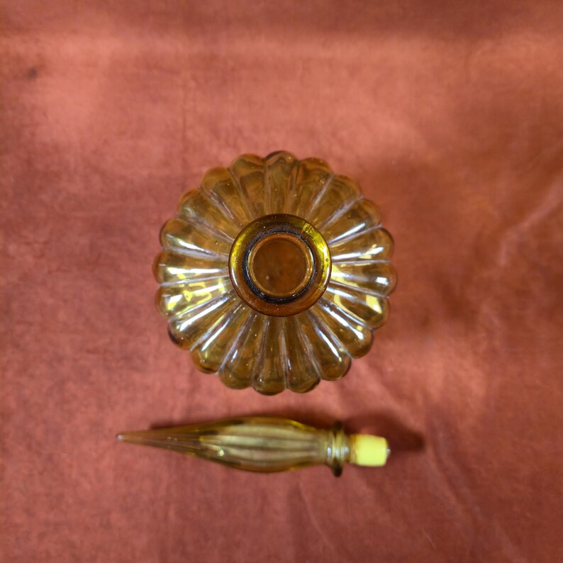 May include: Amber glass perfume bottle with a ribbed, rounded body and a matching stopper. The bottle has a decorative, flower-like design on the top. The stopper has a yellow tip. The bottle is empty.