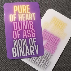 Może przedstawiać: Dwa naklejki z tekstem "PURE OF HEART DUMB OF ASS NON OF BINARY" w białym kolorze na fioletowym i białym tle.