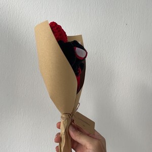 May include: A small bouquet of crocheted flowers in red and black, wrapped in brown paper with a tag that says "CHINO GARDEN".
