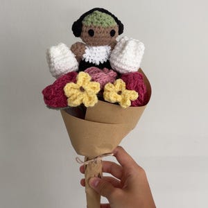 Può includere: Un bouquet di fiori all'uncinetto con una piccola bambola marrone con i capelli verdi e vestita di bianco, circondata da fiori all'uncinetto rosa, bianchi e gialli. Il bouquet è avvolto in carta marrone.