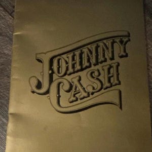 May include: A gold-colored poster with the name "JOHNNY CASH" in a stylized, vintage font. The text is in a dark brown color, with a shadow effect, and is set against a solid gold background. The poster is slightly creased.