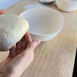 May include: A close-up shot shows a light-colored dough ball being placed into a white, woven bread basket. The basket is round with a textured surface. Another basket is visible in the background, along with a wooden surface.