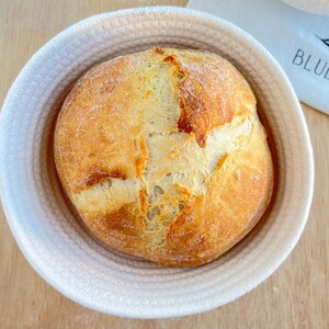 May include: A round loaf of bread with a golden-brown crust sits in a white rope basket. The bread has a cross-shaped score on top. The basket is on a light wooden surface.