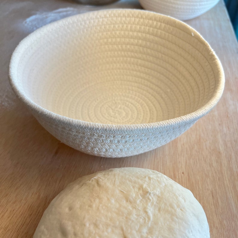 May include: A cream-colored, woven bread proofing basket with a rounded shape. The basket is made of tightly coiled rope, creating a textured surface. A ball of dough sits below the basket on a wooden surface, ready for baking.